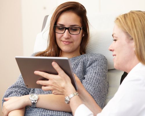 Nach einer Untersuchung der Hautpartien werden einer Patientin die Diagnose und Therapiemöglichkeiten auf einem Tablet vorgestellt. Dabei sitzt die Patientin auf einem Behandlungssessel und die Hautärztin hält das Gerät zur Visualisierung.