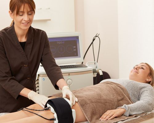 Patientin liegt in ein Handtuch der Hautarztpraxis Heidi Dötterer-Rieg & Kollegen gekleidet auf einer Liege. Eine staatlich geprüfte Kosmetikerin führt eine nicht-chirurgische Fettreduzierung und Figur-Formung Behandlung mit Venus Bliss durch.