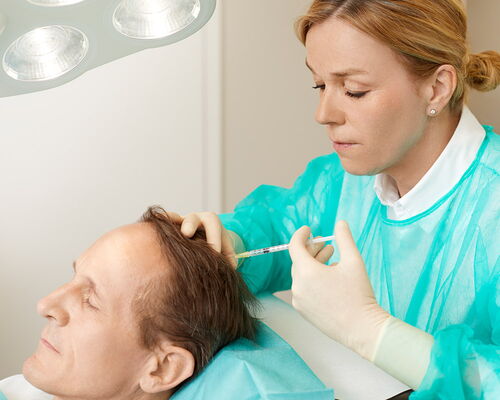 Bei einer Regenera Activa Behandlung gegen erblich bedingten Haarausfall sitzt ein Patient entspannt im Sessel einer hautärztlichen Privatpraxis. Hinter ihm stehend, injiziert ein Hautarzt konzentrierte körpereigene Wachstumsfaktoren in die Kopfhaut.