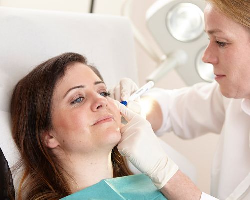 A patient lies relaxed on a couch during a wrinkle treatment. Surgical lights can be seen in the background. Dermatologist at the Frankfurt private practice marks wrinkles with a pen on the patient's face for wrinkle injections.