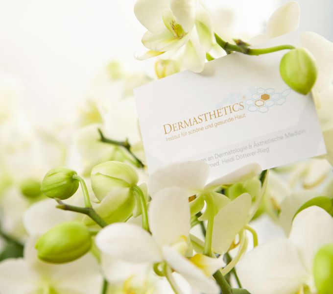 In Mitten von frisch blühenden weißen Orchideen befindet sich eine Visitenkarte des, an die Hautarztpraxis angeschlossenen, medizinisch-kosmetischen dermatologischen Instituts unter fachärztlicher Unterstützung, mit der Bezeichnung Dermasthetics.