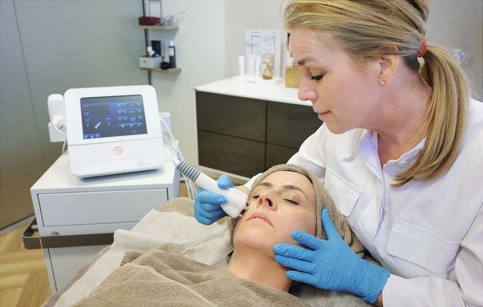 A dermatologist treats the facial skin of a patient in the Frankfurt dermatologist's office. The treated woman's body and head are wrapped in towels. A dermatologist uses a device to massage skin-rejuvenating ingredients into the skin. Device is nearby.