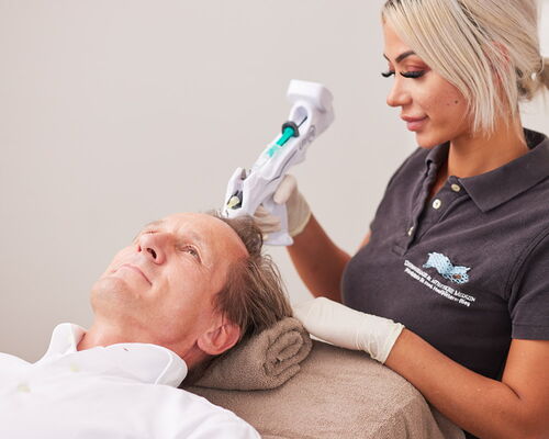 Ein Mann wird mit Mesotherapie gegen Haarausfall behandelt. Er sitzt entspannt in einem Sessel der Privatpraxis für Dermatologie in Frankfurt am Main, während eine medizinische Fachangestellte die Haarwurzel mit der Mesopistole am Haaransatz behandelt.