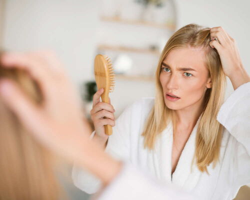 Junge Frau im Bademantel betrachtet sich im Spiegel. In einer Hand hält Sie den Kamm, mit der anderen zieht sie das lange blonde Haar zurück, um den Haaransatz nach einer RPR-Therapie gegen Haarausfall genauer zu betrachten. Ging der Haarausfall zurück?