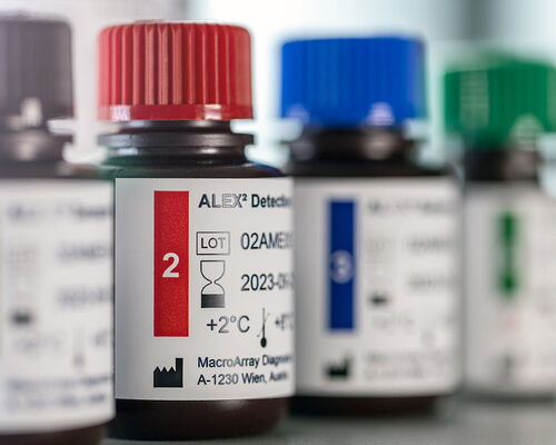 Vier Behälter mit Komponenten für den ALEX Allergie-Test zur Laboranalyse von Allergenen, die die Symptome einer Allergie verursachen. Die Fläschchen für Allergene-Tests stehen in einer Reihe und haben braune, rote, blaue und grüne Verschlusskappen.