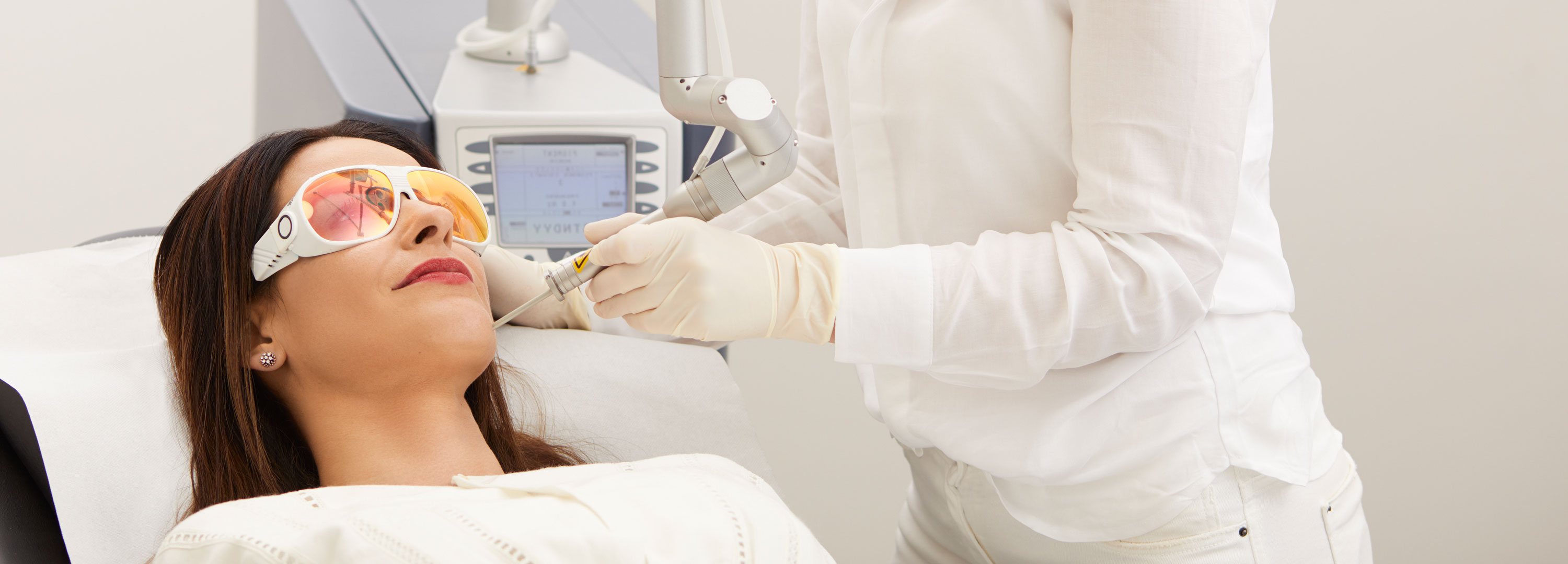 Patientin liegt während einer medizinischen Laserbehandlung auf einer Liege in der Frankfurter Hautarztpraxis. Während der Laser-Hautbehandlung sind ihre Augen mit einer Brille geschützt. Daneben steht eine Fachärztin, die mit modernsten Laser behandelt.