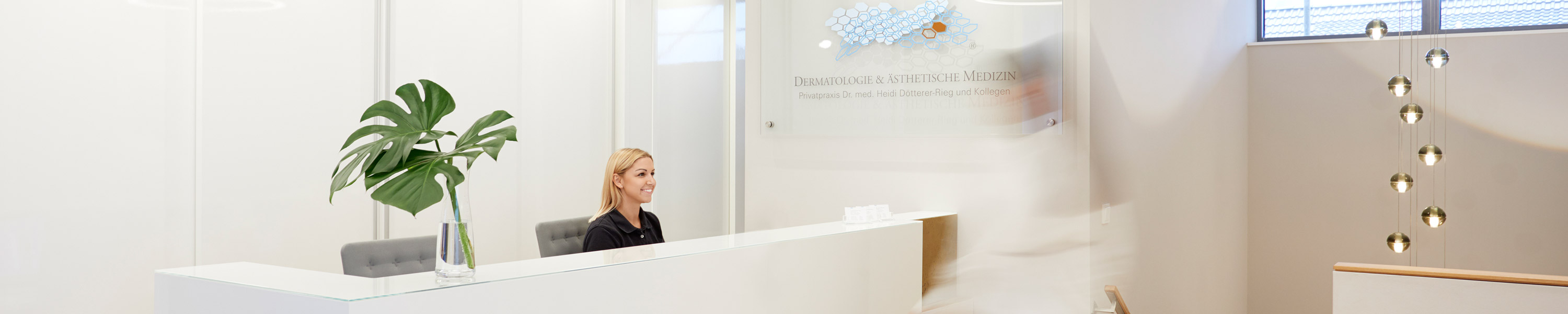A medical assistant receives a patient at the reception desk in the private dermatology practice. The employee of the private medical practice smiles at the patient. The rooms are bright and modernly furnished.